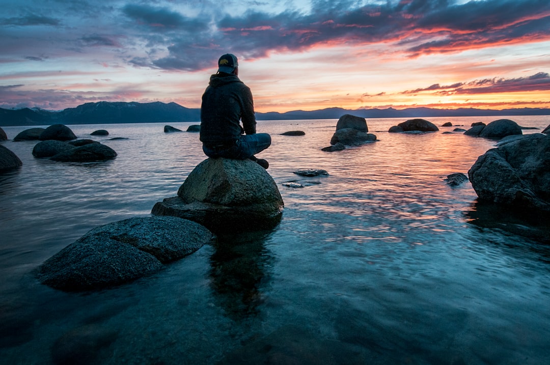 Una persona se sienta en una gran roca en aguas poco profundas, practicando meditación rápida mientras contempla una vívida puesta de sol.