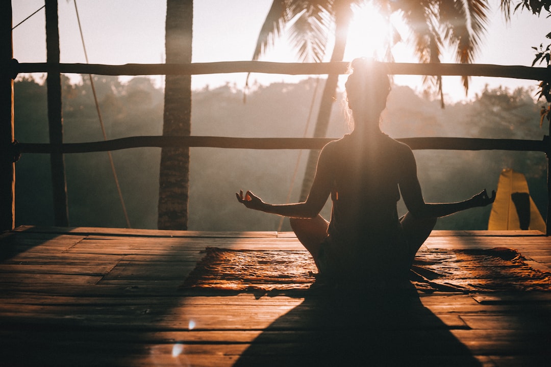 Persona sentada con las piernas cruzadas sobre una esterilla meditando al aire libre al amanecer, con palmeras y la luz del sol de fondo.